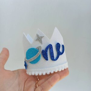 Puede incluir: Una corona de fieltro blanco con un 'uno' de purpurina azul y plata y un planeta azul con un anillo plateado. La corona tiene un ribete de pomp&oacute;n blanco.