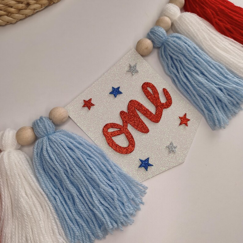 May include: A decorative banner featuring a white glittery pennant with the word one in red script, accented by small red, blue, and silver glitter stars. The banner is strung with natural wooden beads and alternating white, light blue, and red yarn tassels.
