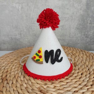 Op de afbeelding: Witte, kegelvormige feestmuts met een rode pompon bovenop. De muts heeft een rode en witte rand en een zwarte glitter 'een' met een pizzapunt-afbeelding.