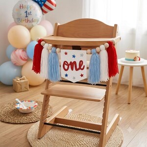 May include: A wooden high chair decorated with a white felt banner featuring the red glitter text 'one' and blue and silver stars. The banner is flanked by red, white, and light blue yarn tassels strung on a wooden bead garland across the front.