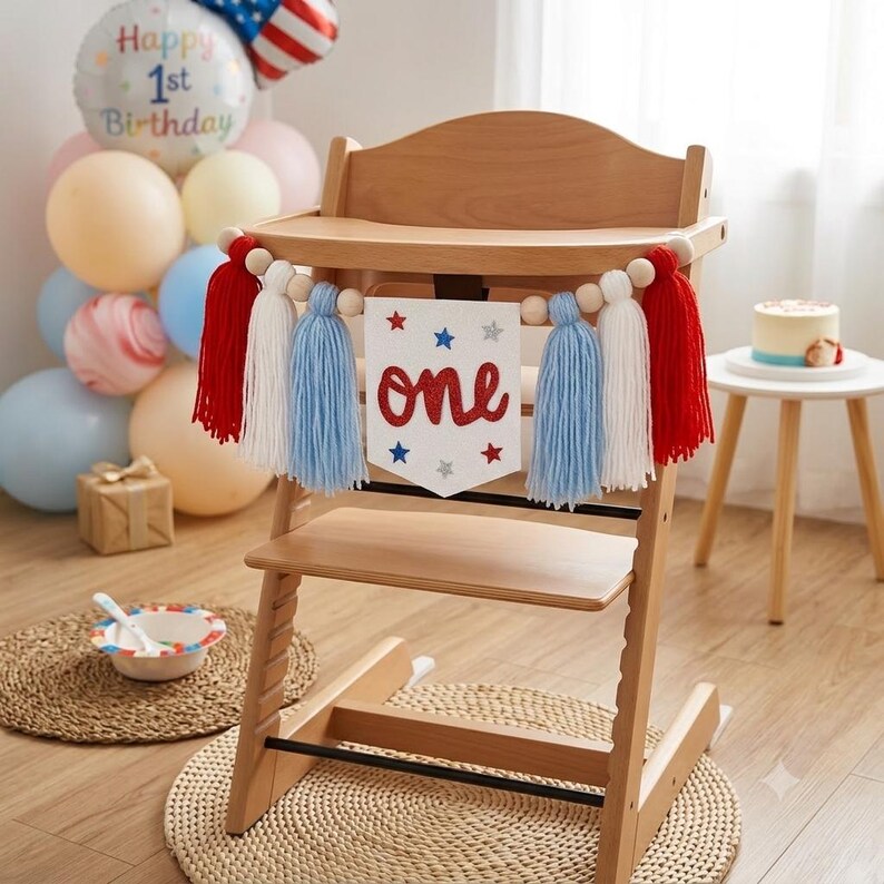May include: A wooden high chair decorated with a white felt banner featuring the red glitter text 'one' and blue and silver stars. The banner is flanked by red, white, and light blue yarn tassels strung on a wooden bead garland across the front.