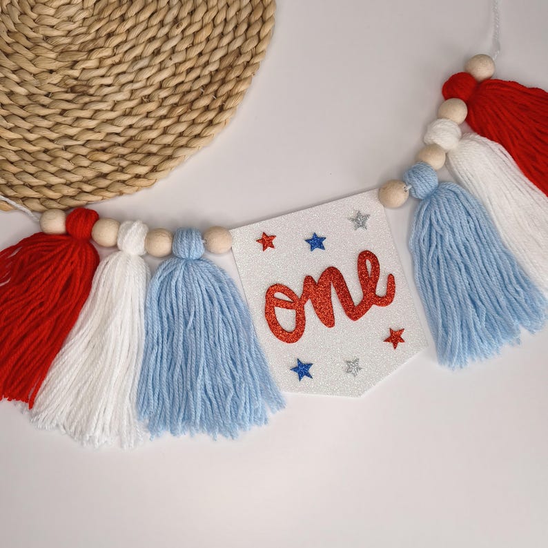 May include: A decorative banner featuring a white glittery pennant with the word one in red script, surrounded by small red, blue, and silver glitter stars. The banner is strung with natural wooden beads and alternating red, white, and light blue yarn tassels.
