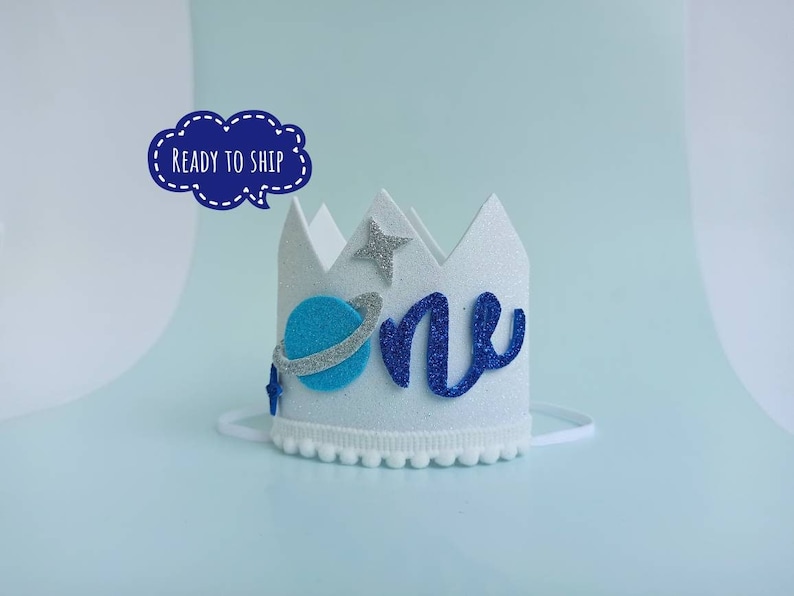 Puede incluir: Corona de fieltro blanco con la inscripci&oacute;n 'uno' en purpurina azul, un planeta azul con un anillo plateado y estrellas plateadas de purpurina. La corona tiene una banda el&aacute;stica blanca.