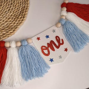 Può includere: Uno stendardo decorativo con un gagliardetto bianco glitterato che riporta la scritta one in corsivo rosso glitterato, circondato da piccole stelle rosse, blu e argento. Il gagliardetto è infilato con perline di legno e nappe di lana colorate.