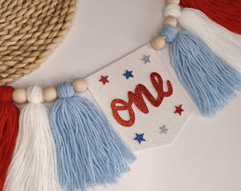 May include: A patriotic banner with red, white, and blue tassels and wooden beads. The banner has a white background with the word "one" in red glitter and star accents, perfect for a first birthday celebration.