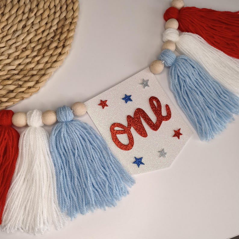 May include: A decorative banner featuring a white glittery pennant with the word one in red glitter script, surrounded by small red, blue, and silver glitter stars. The pennant is strung with natural wooden beads and red, white, and blue yarn tassels.
