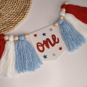 May include: A decorative banner featuring a white glittery pennant with the word one in red script, accented by small red, blue, and silver glitter stars. The banner is strung with natural wooden beads and alternating red, white, and blue yarn tassels.