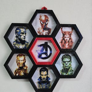 May include: A black and red framed wall art piece featuring portraits of Marvel superheroes. The hexagonal frames surround images of Captain America, Iron Man, Spider-Man, Scarlet Witch, Hulk, Doctor Strange, and the Avengers logo.