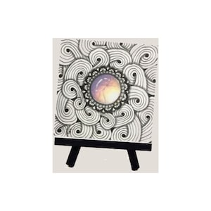 Puede incluir: Una pequeña pintura sobre lienzo en blanco y negro con un patrón de espiral. El centro de la pintura presenta una piedra preciosa redonda, púrpura, rosa y naranja.