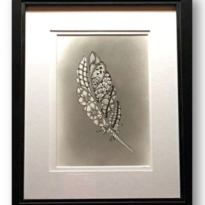 May include: Framed artwork with a black frame, showcasing a detailed grayscale drawing of a feather. The feather is decorated with floral and geometric patterns. It's matted with a white border and set against a gradient grey background.
