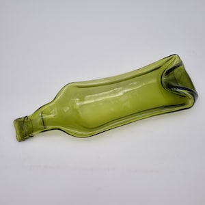 Könnte beinhalten: Eine abgeflachte, olivgrüne Glasflasche, die zu einer Servierschale umfunktioniert wurde. Die ursprüngliche Form der Flasche ist erkennbar, mit Hals und Boden, aber der Körper ist zu einer flachen, rechteckigen Form geformt. Das Glas hat eine leichte Textur und einen subtilen Glanz.