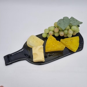 Könnte beinhalten: Ein schwarzes, flaschenförmiges Serviertablett mit Käse und grünen Trauben. Der Käse ist gelb und weiß, mit unterschiedlichen Texturen. Die Trauben sind grün mit ein paar rosa-bräunlichen Trauben.