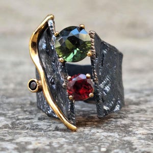 May include: A unique ring featuring a dark gray textured band with gold accents. The ring holds a green gemstone and a red gemstone. The ring's design is artistic and eye-catching, suitable for a special occasion or everyday wear.