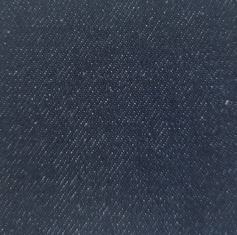 May include: Close-up of dark blue denim fabric. The fabric has a diagonal weave pattern with small, white flecks scattered throughout. The texture appears slightly rough, typical of denim.