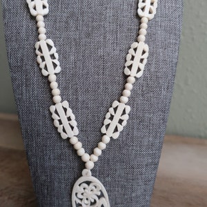 Op de afbeelding: Een witte, gesneden ketting van been met een grote ovale hanger. De hanger heeft een bloemmotief.