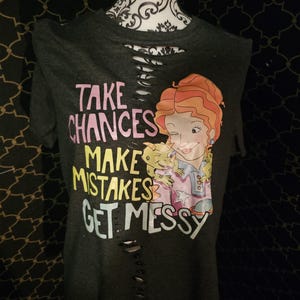 Könnte beinhalten: Ein dunkelgraues T-Shirt mit einer Grafik einer Frau mit orangefarbenen Haaren und einer kleinen grünen Kreatur. Der Text lautet "TAKE CHANCES MAKE MISTAKES GET MESSY". Das Shirt hat eine Reihe horizontaler Schnitte im Brustbereich.