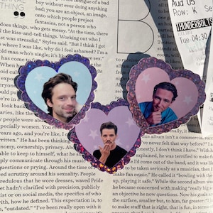May include: Three heart-shaped stickers with images of men's faces, set against a backdrop of printed text. The stickers have a glittery border and a light blue background with star accents.