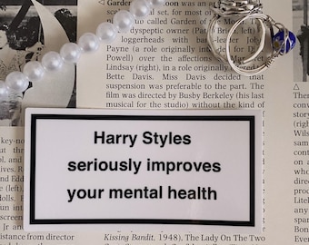 May include: A pearl necklace and a silver ring with a rose design and a blue bead rest on a newspaper background. A sticker with the text "Harry Styles seriously improves your mental health" is also visible.