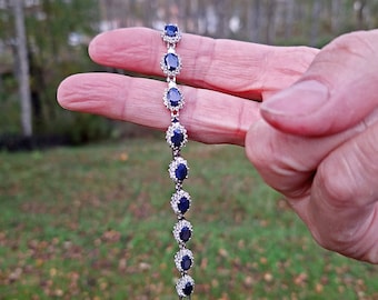 May include: A silver bracelet featuring oval, dark blue gemstones surrounded by smaller, clear stones. The bracelet is held up against a blurred green and brown background, showcasing its elegant design and color contrast.