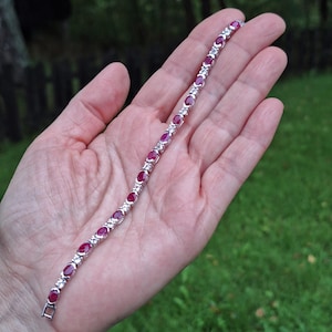 Puede incluir: Un brazalete plateado con piedras preciosas rojas oscuras de forma ovalada, alternando con piedras más pequeñas y transparentes. El brazalete se muestra sobre un fondo verde, resaltando su diseño elegante y su color vibrante.