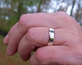 May include: A silver ring with a polished finish. The ring is a simple band design, reflecting light. The ring is on a finger, with a blurred background of green and brown.