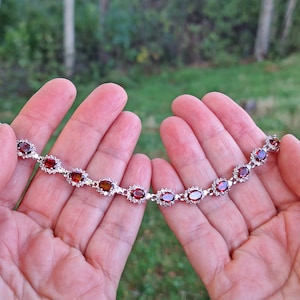 Può includere: Un bracciale in argento con pietre preziose ovali rosso scuro, ciascuna circondata da pietre più piccole e trasparenti. Il bracciale è mostrato su uno sfondo verde sfocato, evidenziando la lucentezza e il design del gioiello.