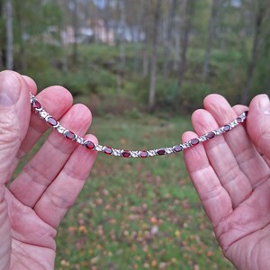 May include: A silver bracelet featuring alternating oval-shaped, deep red gemstones and clear, round-cut stones. The bracelet is held up against a blurred green and brown background, showcasing its delicate design and vibrant colors.