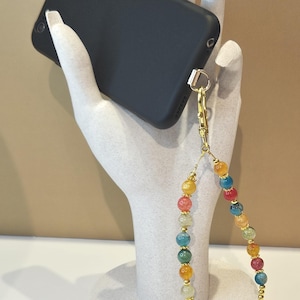 May include: A black smartphone case attached to a beaded wrist strap. The strap features multicolored, crackled-texture round beads in shades of red, yellow, blue, green, and orange, interspersed with small gold-toned metal spacer beads and a gold clasp.