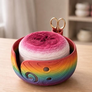 May include: A rainbow-colored yarn bowl holds a large ball of pink and white yarn. The bowl has a curved design with a slot for the yarn and a holder for gold scissors. The bowl sits on a wooden surface.