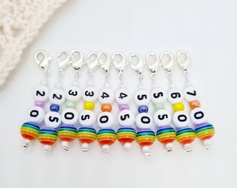 May include: A collection of ten stitch markers with silver clasps. Each marker features a white bead with a number, and a rainbow-striped bead. The numbers range from 20 to 70. The markers are designed for knitting or crochet projects.