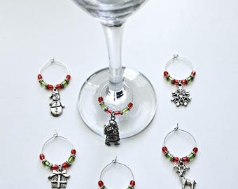 May include: A collection of Christmas-themed wine charms. Each charm features a silver-toned pendant, including a snowman, snowflake, gift, Santa, Christmas tree, and reindeer. The charms are adorned with red and green beads, attached to a silver wire ring.