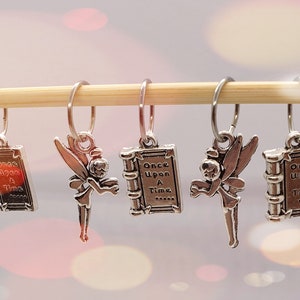 May include: Five silver charms hang from a wooden dowel. The charms feature a fairy and a book with the words "Once Upon A Time" engraved. The charms are attached to silver hoops.