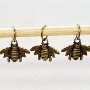 May include: Five small, bronze-colored bee charms with detailed wings and bodies hang from small hoops. They are suspended from a light-colored wooden dowel against a white background. The bee charms are evenly spaced.