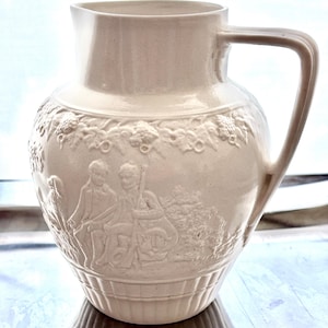 May include: A cream-colored ceramic pitcher featuring a relief design of two figures sitting outdoors with a dog. The upper section displays a grape vine motif, while the base is decorated with vertical fluting. It has a handle and a spout.