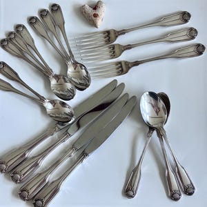 May include: A collection of silver-toned flatware including three dinner knives, three dinner forks, and six spoons. Each piece features a decorative shell motif at the base of the handle. A small, heart-shaped textured stone sits among the utensils.