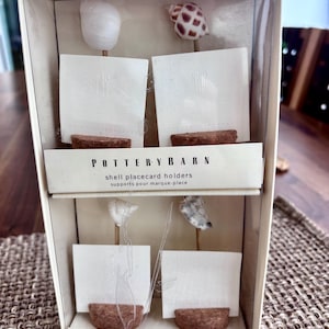 May include: A Pottery Barn set of four shell placecard holders in a clear box. Each holder features a natural seashell topper on a thin stem, anchored by a semi-circular cork base. The set includes four white square cards with a subtle utensil icon.
