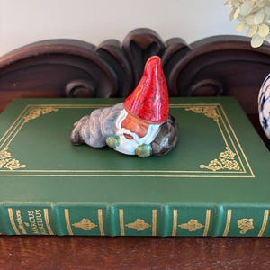 May include: A ceramic figurine of a sleeping gnome wearing a bright red pointed hat with black speckles, a gray robe, and a white beard. The gnome is resting on a green hardcover book titled Meditations of Marcus Aurelius with gold accents.