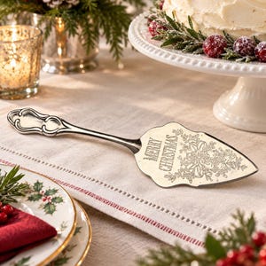May include: A silver-toned metal cake server with an ornate handle and a triangular blade. The blade features an engraved design of a bow and holly leaves, along with the text "MERRY CHRISTMAS" etched into the surface in a decorative font.