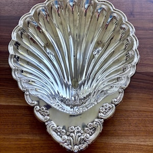 May include: A silver-toned metal dish shaped like a large scallop shell. The interior features deep, radiating fluted grooves. The base of the shell is decorated with an ornate, raised floral relief pattern featuring small blossoms and scrolling leaves.