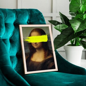 May include: A framed print of the Mona Lisa with a yellow paint stroke covering her eyes. The print is sitting on a teal velvet armchair.