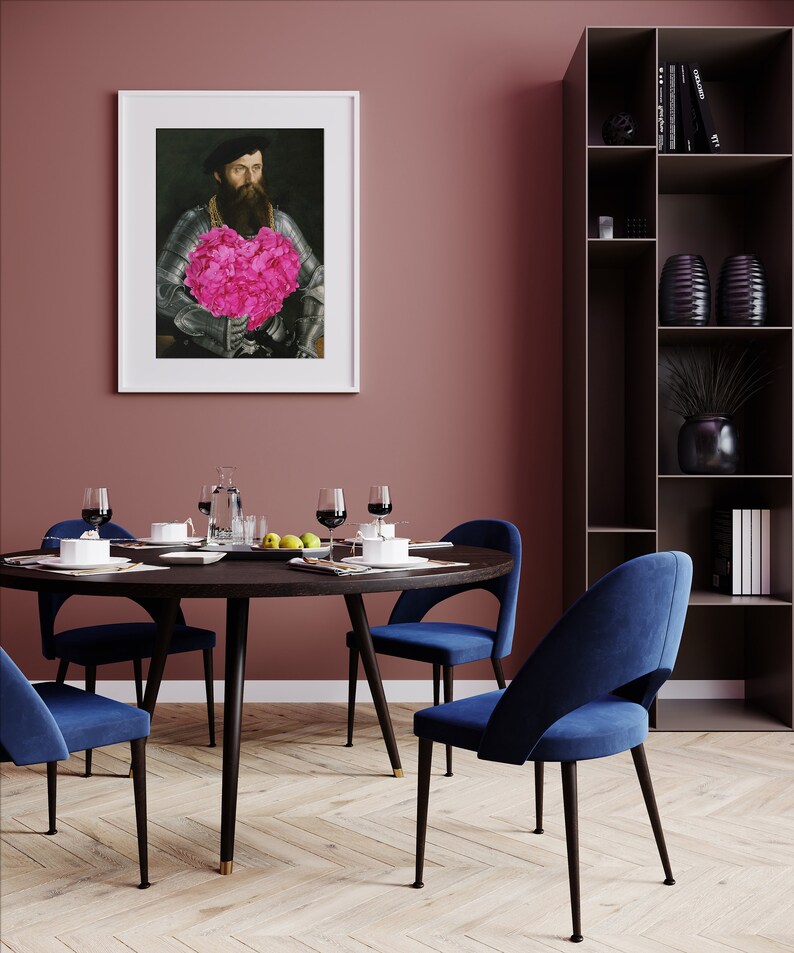 May include: A round dining table with a dark wood top and black metal legs. Four blue velvet chairs surround the table. A framed portrait of a man holding a pink flower bouquet hangs on the wall behind the table. A tall black bookshelf with shelves filled with books and decorative objects is in the background.