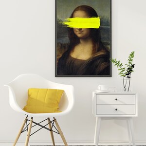 May include: A black framed print of the Mona Lisa painting with a yellow paint stroke covering her eyes. The print is hanging on a white wall above a white chair with a yellow pillow and a white nightstand with two drawers.
