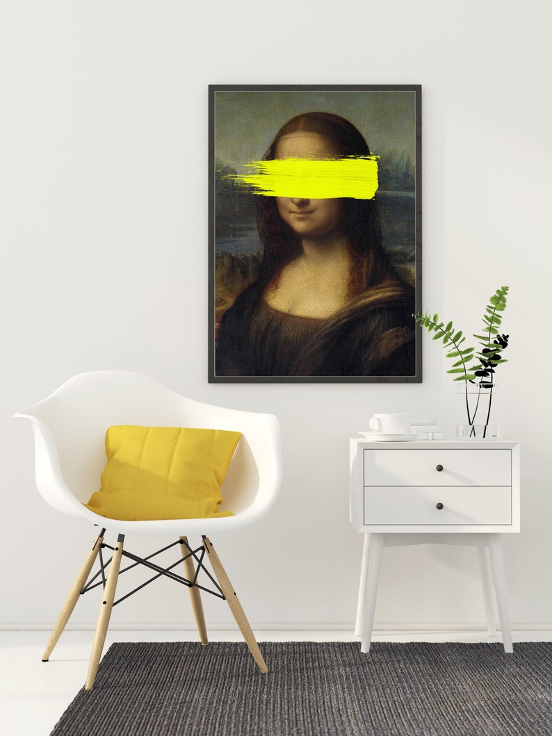 May include: A black framed print of the Mona Lisa painting with a yellow paint stroke covering her eyes. The print is hanging on a white wall above a white chair with a yellow pillow and a white nightstand with two drawers.