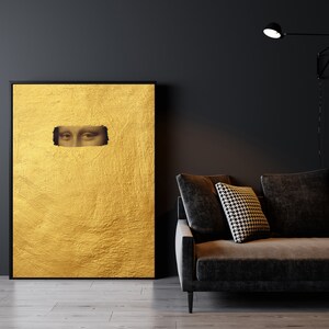 May include: A framed artwork featuring a textured gold background with a rectangular cutout revealing the eyes of the Mona Lisa. The artwork is displayed against a dark wall, next to a dark gray sofa with a patterned pillow.
