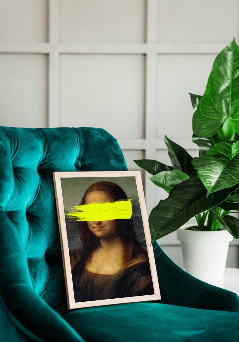 May include: A framed print of the Mona Lisa with a yellow paint stroke covering her eyes. The print is sitting on a teal velvet armchair.