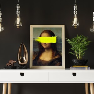 May include: A white desk with two drawers and wooden legs. A framed print of the Mona Lisa with a yellow paint stroke covering her eyes sits on the desk. A small wooden elephant figurine and a gold metal abstract sculpture are also on the desk. A potted green plant and three hanging light bulbs are in the background.