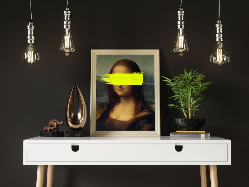 May include: A white desk with two drawers and wooden legs. A framed print of the Mona Lisa with a yellow paint stroke covering her eyes sits on the desk. A small wooden elephant figurine and a gold metal abstract sculpture are also on the desk. A potted green plant and three hanging light bulbs are in the background.