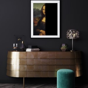 May include: A framed print of the Mona Lisa, partially obscured by a black vertical stripe, hangs above a gold-toned cabinet. A green velvet stool and a crystal lamp add to the room's decor. The room has dark walls and a wooden floor.