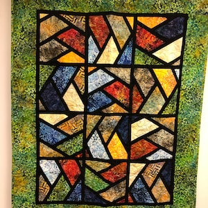 Puede incluir: Una colcha con un diseño de vitral que presenta formas geométricas en tonos de azul, rojo, amarillo y verde. La colcha está enmarcada con un borde verde.