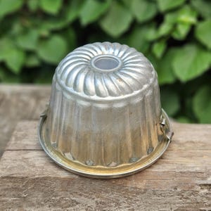 Könnte beinhalten: Eine silberfarbene Bundt-Kuchenform mit geriffeltem Design. Die Form hat einen kreisförmigen Boden und ein zentrales Rohr. Die Metallform weist Gebrauchsspuren auf, was darauf hindeutet, dass es sich um einen Vintage-Küchenartikel handelt.
